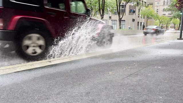 A low angle handheld slow motion view of cars splashing water onto the sidewalk after a heavy rain in a large city.  	