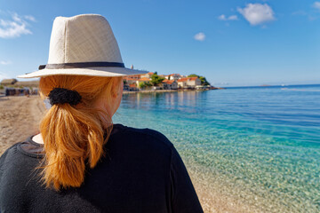 Red head, young woman with red hair and white sun hat stands on the beach and looks at the Old Town Primosten in Croatia. The water is wonderfully clear and blue.