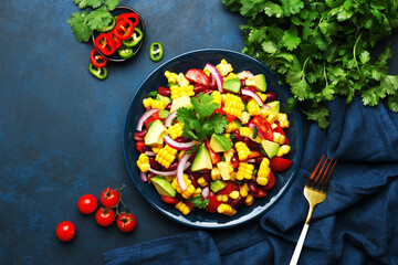 Mexican spicy salad with corn, red beans, avocado, jalapeno peppers, cherry tomatoes, onion and cilantro. Blue table background, top view