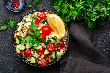 Bulgur tabbouleh salad with fresh tomatoes, cucumbers, parsley and lemon dressing. Traditional Middle Eastern and Arabic dish. Black stone kitchen table background, top view