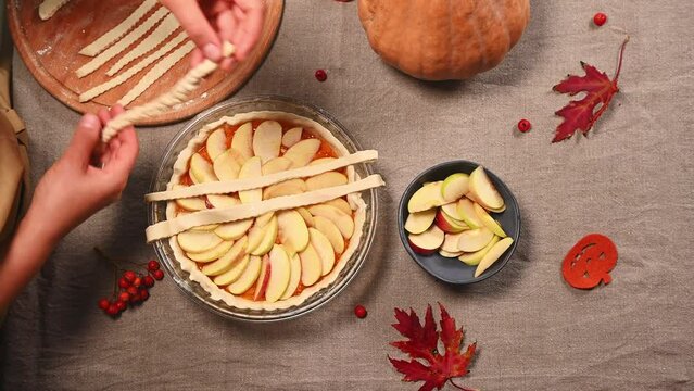 Top view of housewife or chef hands decorating traditional American classic apple pie with pumpkin, by laying sliced pastry strips in lattice pattern, while making homemade Thanksgiving pie