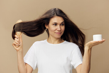 a beautiful, happy, sweet woman stands on a beige background in a white tank top and combs her beautiful, well-groomed, dark, shiny hair with a comb, holding a white cream jar in her hand