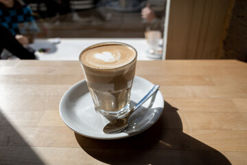 Flat white coffee in clear transparent glass and white plate lay on rough wooden table counter beside windows of coffee shop and blur background of outdoor seats and people outside coffee shop.