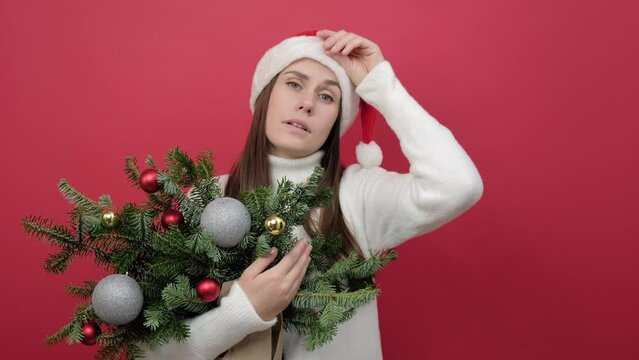Portrait of sick ill young caucasian woman wears white sweater and Santa hat holding bouquet of spruce branches hand on forehead having headache suffer from migraine, isolated on red background studio