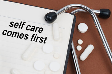 On a brown surface lies a stethoscope, pills and a notepad with the inscription - self care comes...