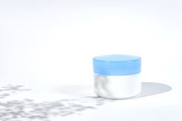 Cosmetic white jar blue lid with cream or lottion and flowers shadows. White background with daylight and harsh shadows