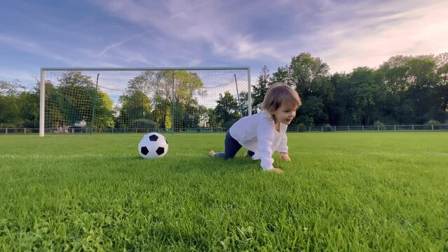 Little kid plays football at sunset, baby runs around green football field and kicks ball, girl runs to floor in meadow and smiles, learns how to kick ball, dream of becoming team footballer