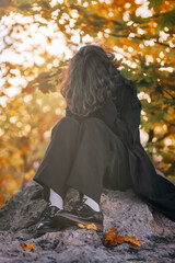 portrait of curly-haired teenage girl in black raincoat sitting on stone in autumn maple forest,...