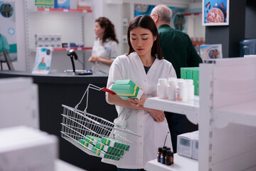 Pharmacist woman gathering pills packages, putting in basket to rearrange pharmacy shelves. In...