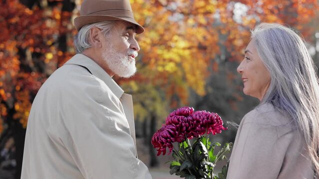 Happy old couple walking holding hands, talking and smiling to each other. Middle aged spouses enjoying time together in beautiful autumn park. Flowers, romantic relationship and dating concept.
