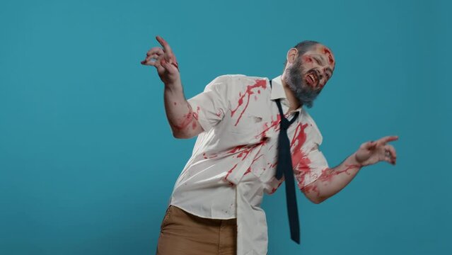 Goofy ugly zombie dancing childish on blue background. Creepy mindless brain-eating monster with deep and bloody wounds and scars enjoying dance while looking at camera. Studio shot