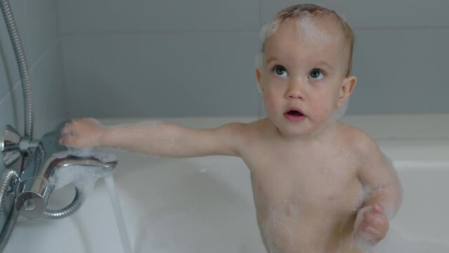 little naked baby child standing in bath tube filling hot water soap foam holding water tub turning on and off water flow. caucasian baby toddler having bath with foam for the first time baby emotions