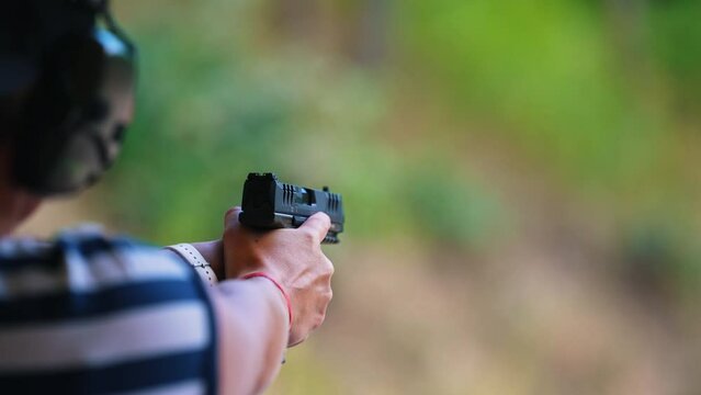Outdoor shooting range concept. Back view of an unrecognizable caucasian person tightly holding basic black handgun, aiming, and shooting at target. Flying shell. Blurred background. High quality 4k