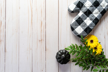 Flat lay with a black and white oven mitt, sunflowers, leaves and a black pumpkin on a light wood...