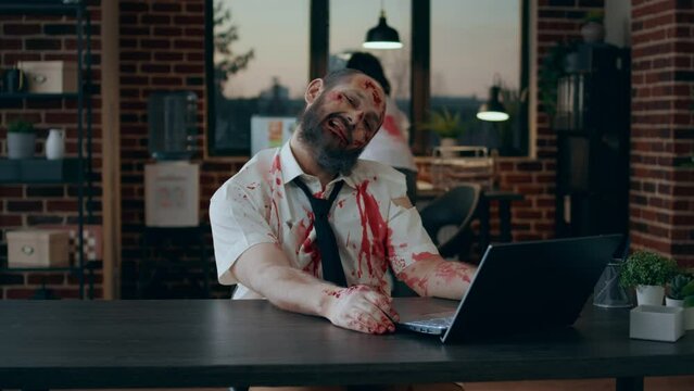 Mindless brain-eating zombie working on laptop while smirking creepy at camera. Dangerous brain dead monster with deep and bloody scars trying to use computer but fails big time.
