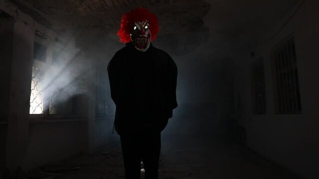 Scary evil clown with red hair standing inside ruined hall in abandoned building at night. Selective focus