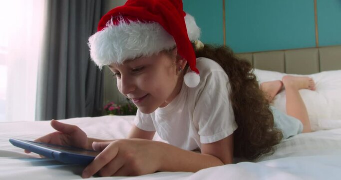 Curious happy cute smiling child in Christmas hat Santa Clausa, kid girl using digital tablet technology device lying on bedroom alone. Small child hold pad computer surfing internet play game at home