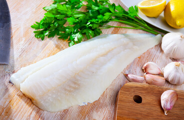 Fresh raw cod fillet lying on wooden cutting board with greens and spices