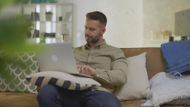 Businessman working from home on laptop computer. Young man sitting on couch in home office with laptop, thinking.