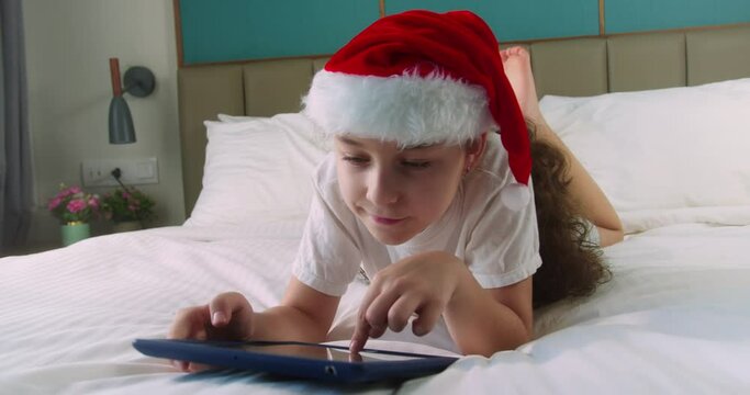 Happy cute smiling child in Christmas hat Santa Clausa, kid girl using digital tablet technology device lying on bedroom alone. Small child hold pad computer surfing internet play game at home
