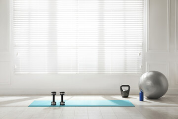 Exercise mat, dumbbells, kettlebell, fitness ball and bottle near window in spacious room