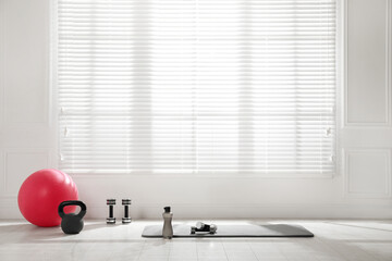 Exercise mat, dumbbells, kettlebell, fitness ball and bottle near window in spacious room