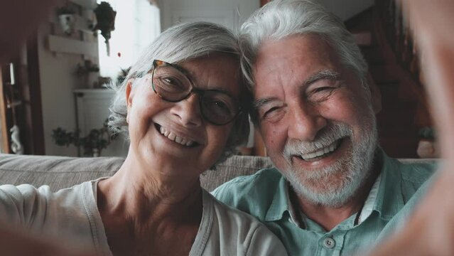 Couple of two old and happy seniors having fun at home on the sofa doing a heart shape with their hands and fingers looking at the camera.