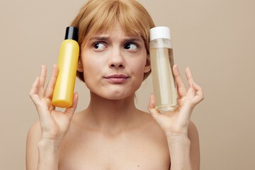 Fototapeta premium a funny, anxious woman stands on a beige background holding jars of care cosmetics in her hands and making an emotional face presses them to her head. Horizontal photo with empty space