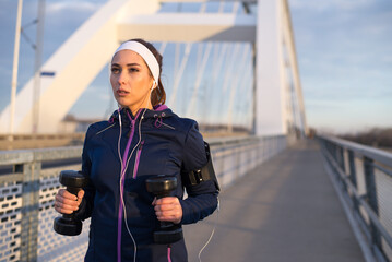 Young sport woman running on the bridge carrying dumbbells on a sunny day