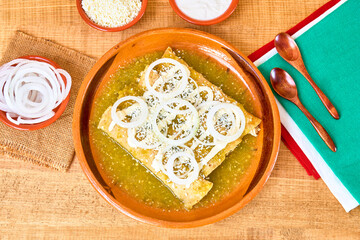 Mexican food (green enchiladas) served with onion rings, aged cheese and cream in a clay dish on a wooden table, decorated with colorful napkins of the Mexican flag. top view