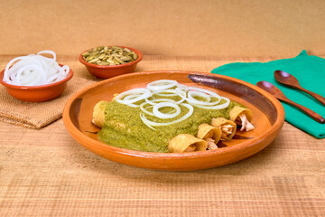Mexican food (green mole enchiladas) served with onion rings in a clay dish on a wooden table, decorated with colorful napkins of the Mexican flag. copy space