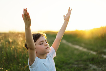 Boy closed her eyes and praying in a field at sunset. Hands folded in prayer concept for faith,...