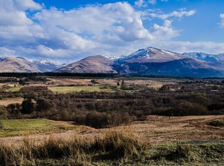 Beautiful farms and mountains, Scottish Highlands, Scotland. Blue sky with clouds, and snow on the peaks in background, farmland with green and brown fields in front. Long view, peaceful scene.