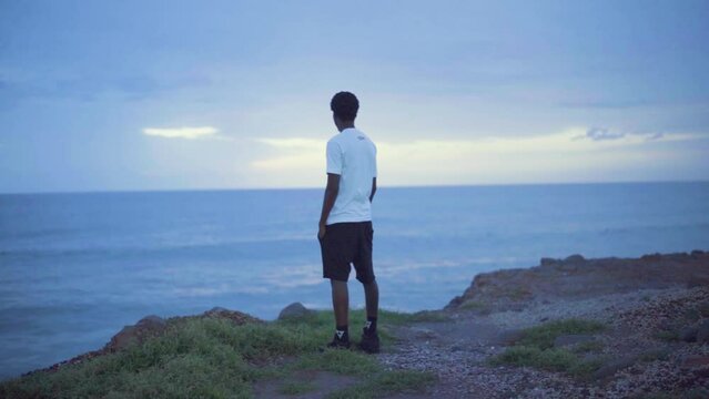 Young black man standing on a cliff looking at the sunset and the ocean.