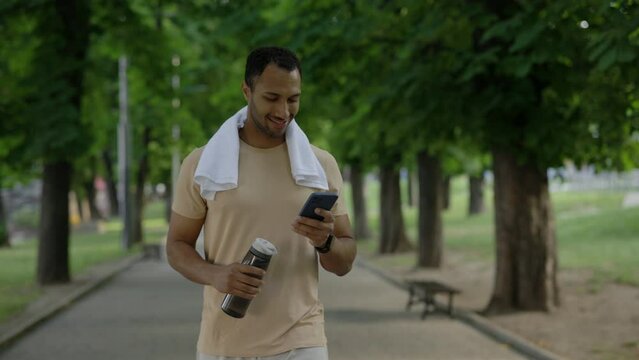 Male african american runner is walking in the park after running using smartphone holding sport bottle and carries a towel around his neck wearing a smart watch on his arm.