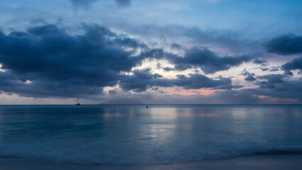 Evening twilight on a tropical island. The turquoise ocean is calm. Blue clouds in the sky, highlighted in pink. Reflection on the water. Foam of waves on a sandy beach. Long exposure. Seychelles. 