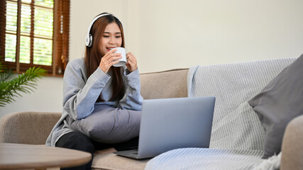 Relaxed Asian female with headphones watching movie on her laptop while sipping coffee