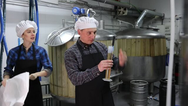 Couple of professional brewmasters standing on background of fermenters at brewery, using test tube and hydrometer to check alcoholic content in craft beer
