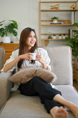 Obraz premium Beautiful Asian female leaning on sofa, sipping coffee while looking at the view out of window