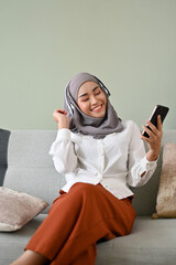 Joyful Asian Muslim woman enjoys listening music through her headphones