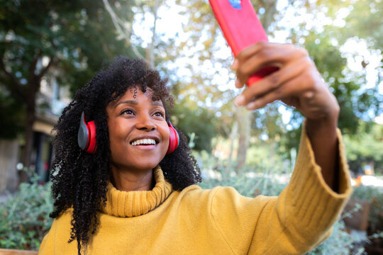 Smiling young African American woman with headphones taking selfie with smartphone in city park