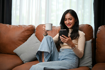 Attractive young Asian female using her smartphone while sitting and sipping coffee on sofa