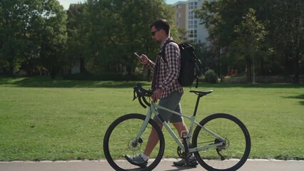 A male cyclist walks his bicycle through a park and uses a smartphone in sunny weather in Germany. A man is pushing a bicycle and surfing the Internet on his phone in a city park in Europe 