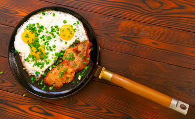 Home made fried eggs and bacon on frying pan with green onion and ground pepper. On wooden table top. Flat lay.