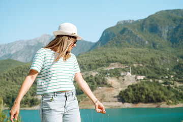 Fototapeta na wymiar Smiling woman in hat and sunglasses with wild hair standing near mountains lake on background. Positive young woman traveling on blue lake outdoors travel adventure vacation.