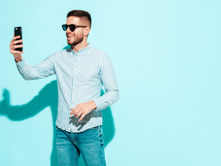 Handsome model. Sexy stylish man dressed in shirt and jeans. Fashion hipster male posing near blue wall in studio. Holding smartphone. Looking at cellphone screen. Using apps. In sunglasses