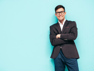Obraz na płótnie Canvas Portrait of handsome smiling model. Sexy stylish man dressed in blue suit. Fashion hipster male posing near blue wall in studio. In glasses. Crossed arms