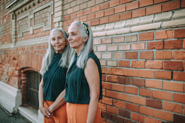 Fototapeta na wymiar Senior women twins,in same clothes standing and posing in front of brick wall, outdoor in city.