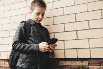 Schoolboy with backpack using smartphone, texting outdoors standing on brick wall urban background. Teenager boy playing video game in internet. Social media
