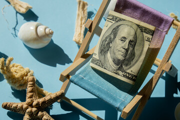 Rolled Up Hundred Dollar Banknote On Miniature Deck beach Chair. Concept of costs in travel...